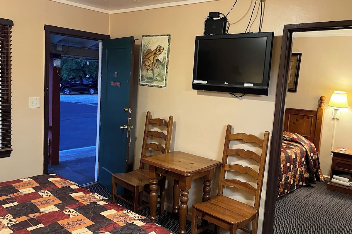 Quarto de hotel com cama, escrivaninha, duas cadeiras, televisão e porta para o exterior.