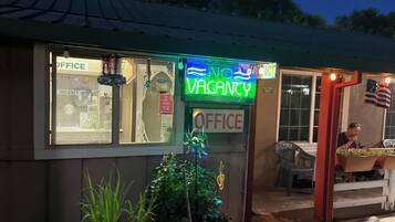 A building with a neon sign indicating 'VACANCY' and an 'OFFICE' sign. There is a chair outside and a plant in the foreground.