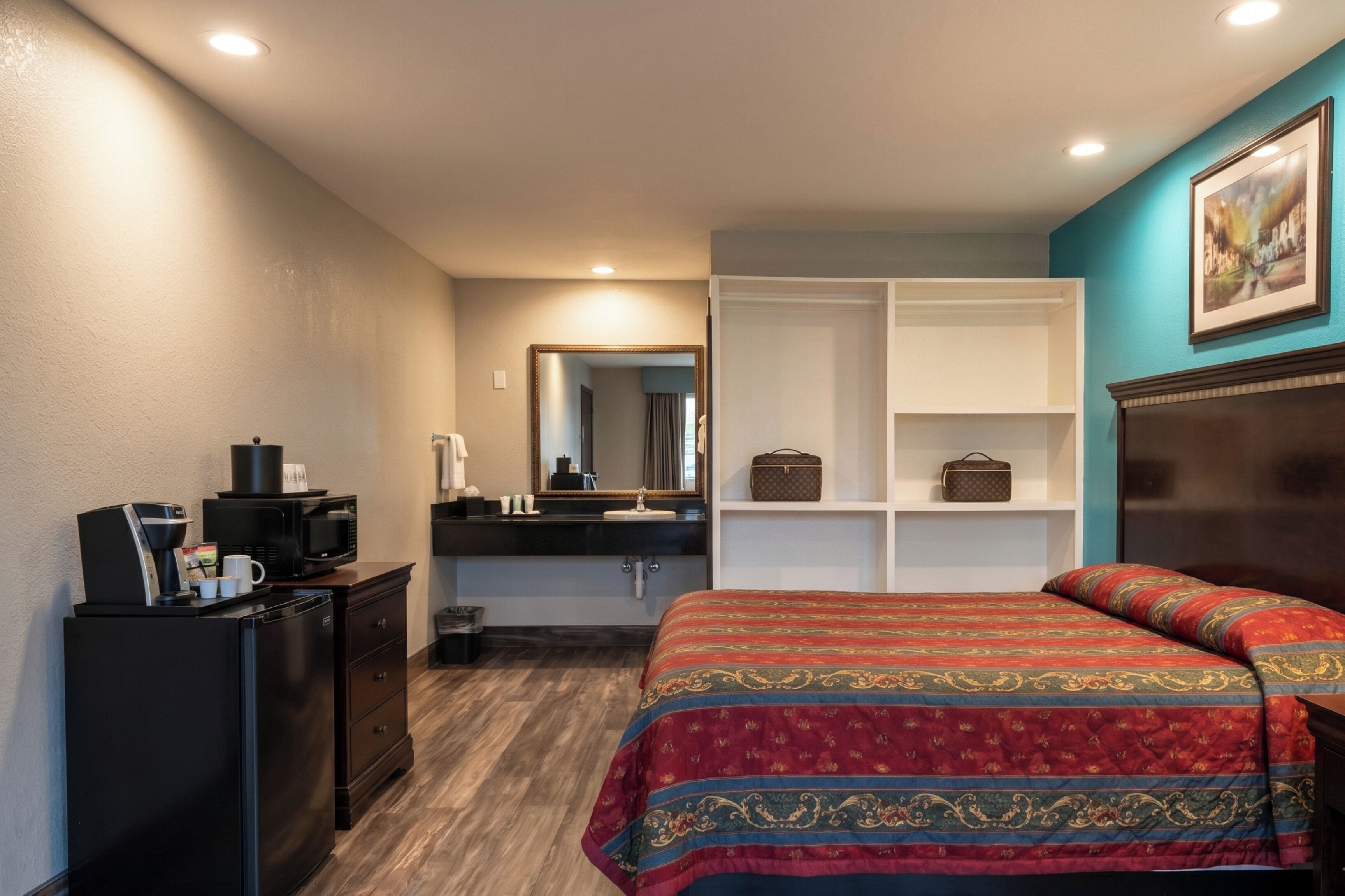 A hotel room with a bed, a dresser, a microwave, a coffee machine, a mirror, and a shelf with a handbag.