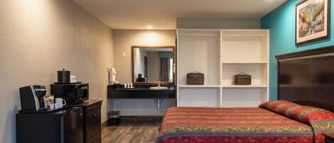 A hotel room with a bed, a dresser, a microwave, a coffee machine, a mirror, and a shelf with a handbag.