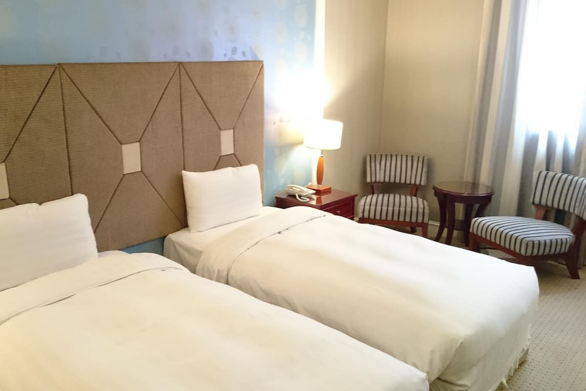 A hotel room with two beds, a headboard with geometric patterns, a bedside table with a lamp, and two striped chairs.