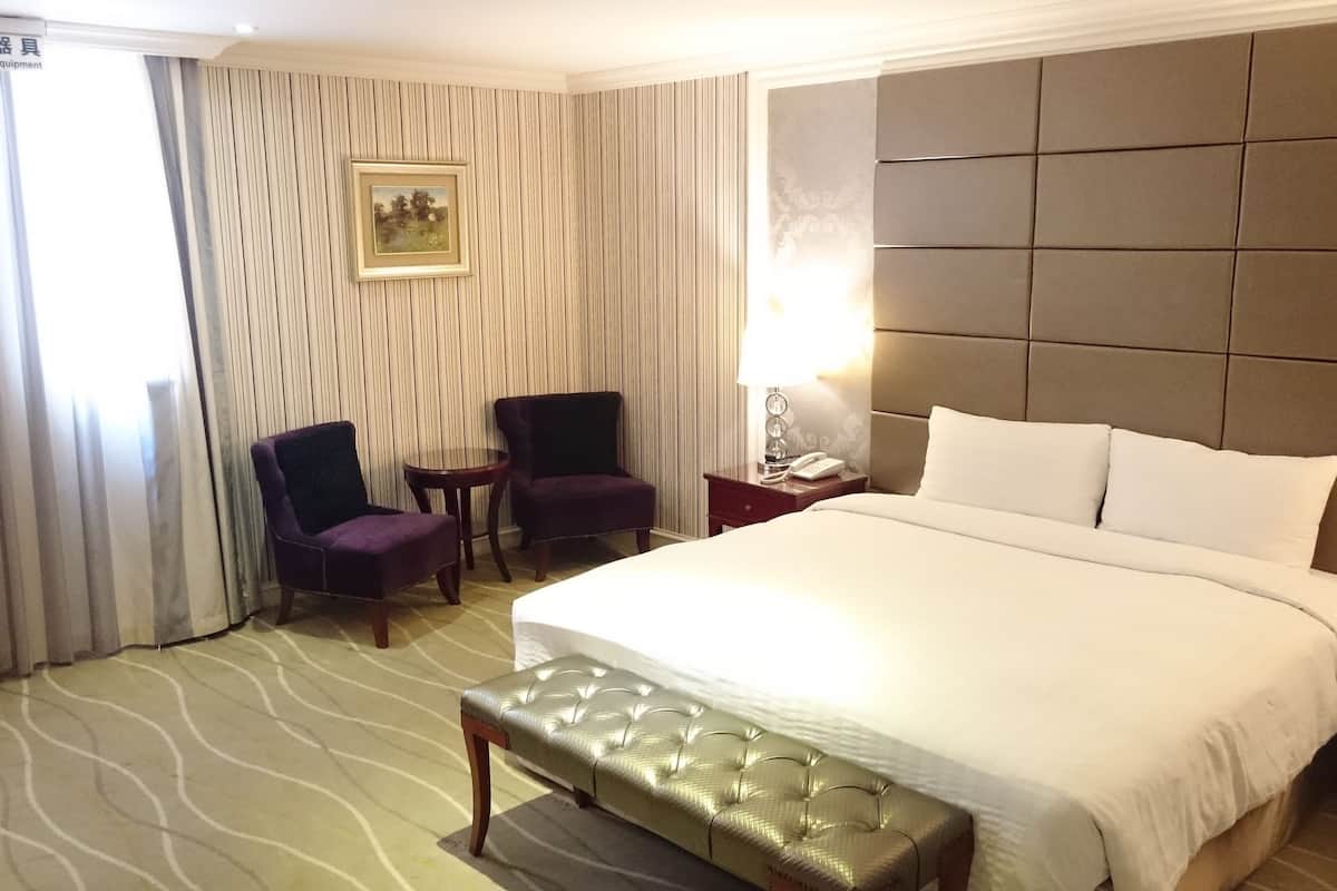 A hotel room with a large bed, a bench, two chairs, a bedside table with a lamp, and a framed picture on the wall.