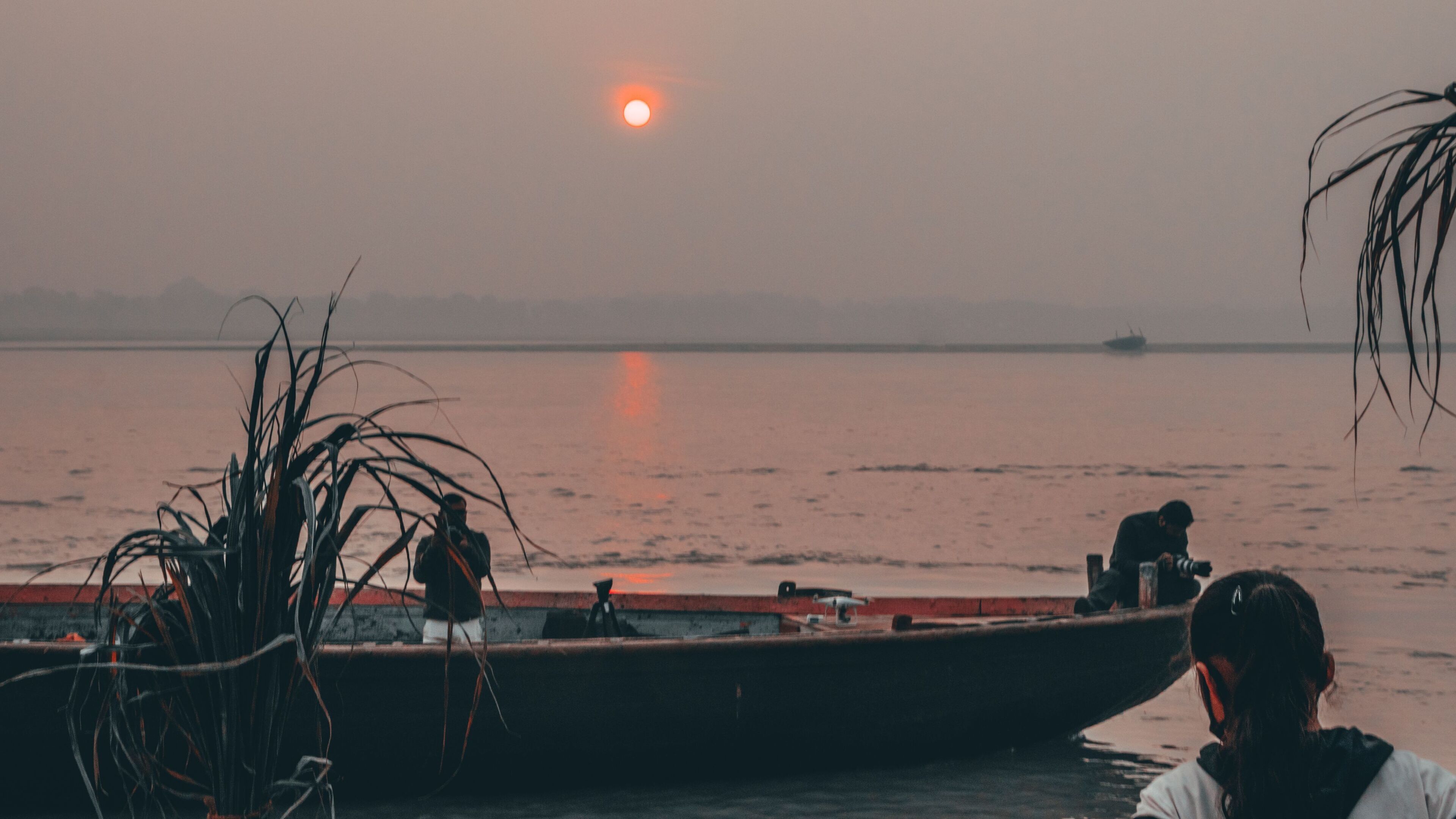 A boat with two people on it, docked at a pier with a calm body of water and a sunset in the background.