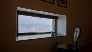 A room with a window overlooking a body of water, a chair, and framed pictures on the wall. - Amritara Suryauday Haveli (Varanasi)