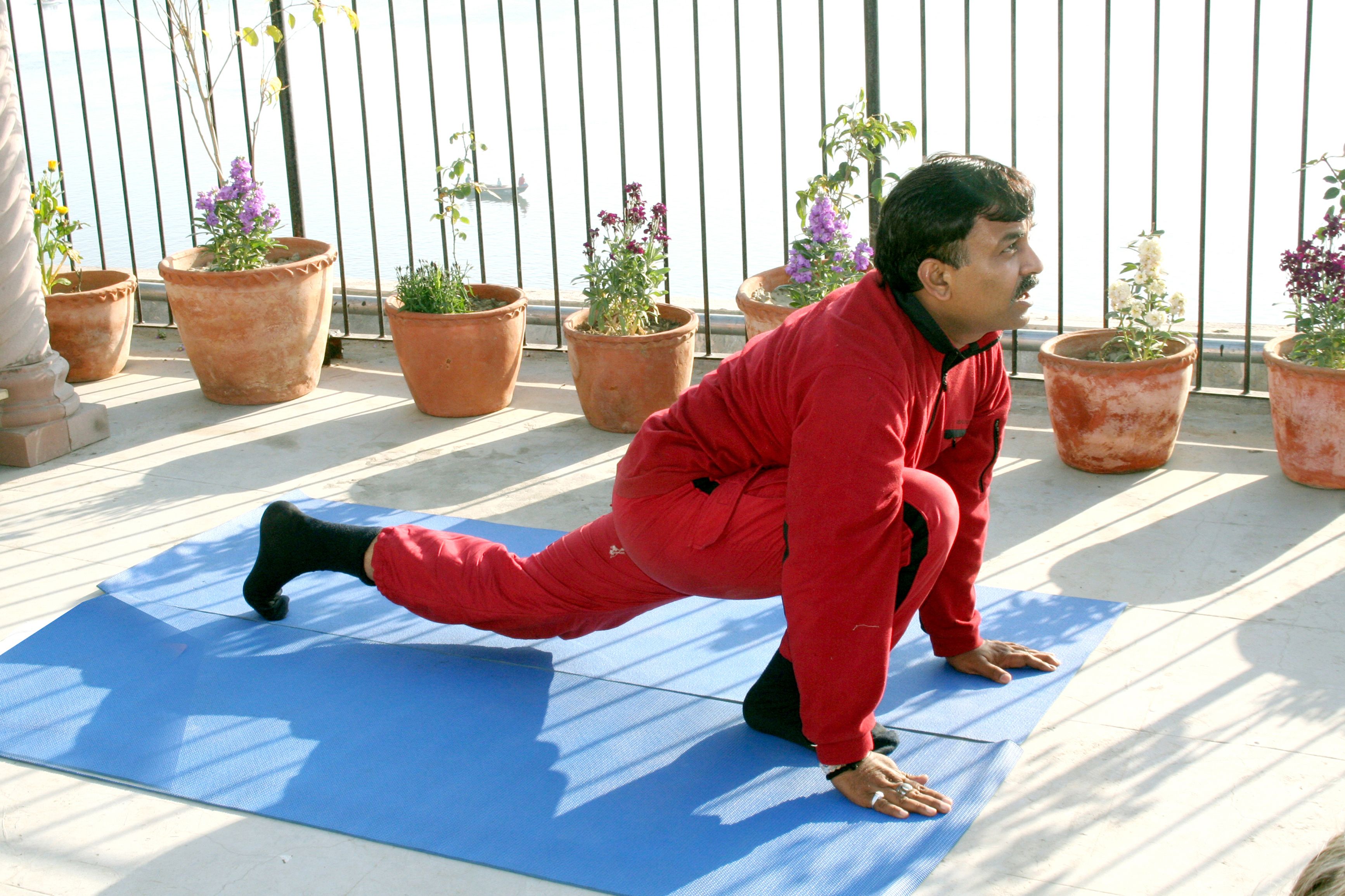 A person in red workout attire performing a yoga pose on a blue mat outdoors, with potted plants and a metal railing in the background.