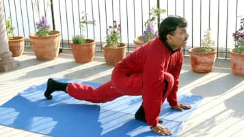 A person in red workout attire performing a yoga pose on a blue mat outdoors, with potted plants and a metal railing in the background.