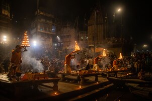 A nighttime outdoor event with people performing rituals by a river, surrounded by buildings.