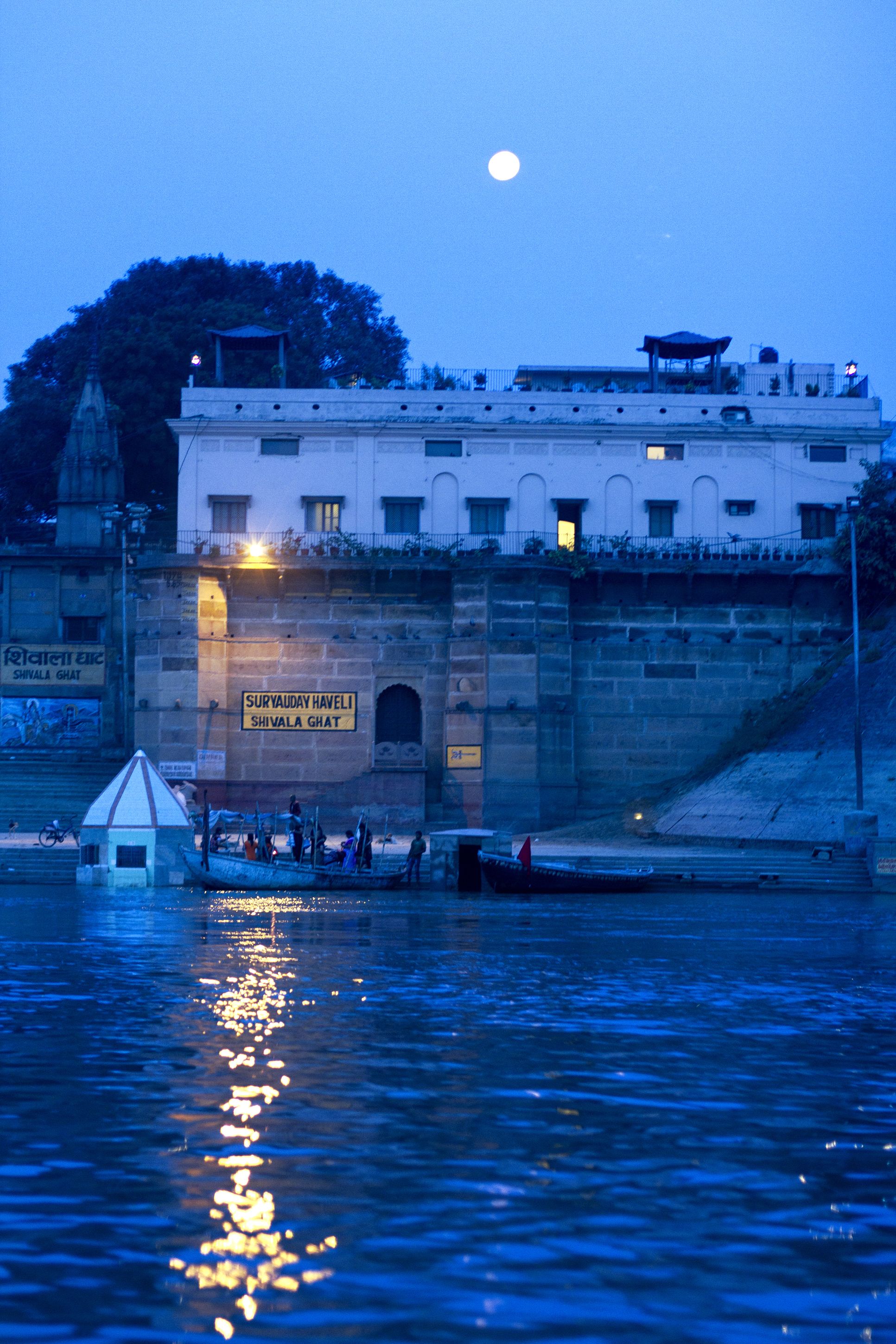 A riverside view with a building featuring a sign reading "Suryaayad Haveli Shivala Ghat".