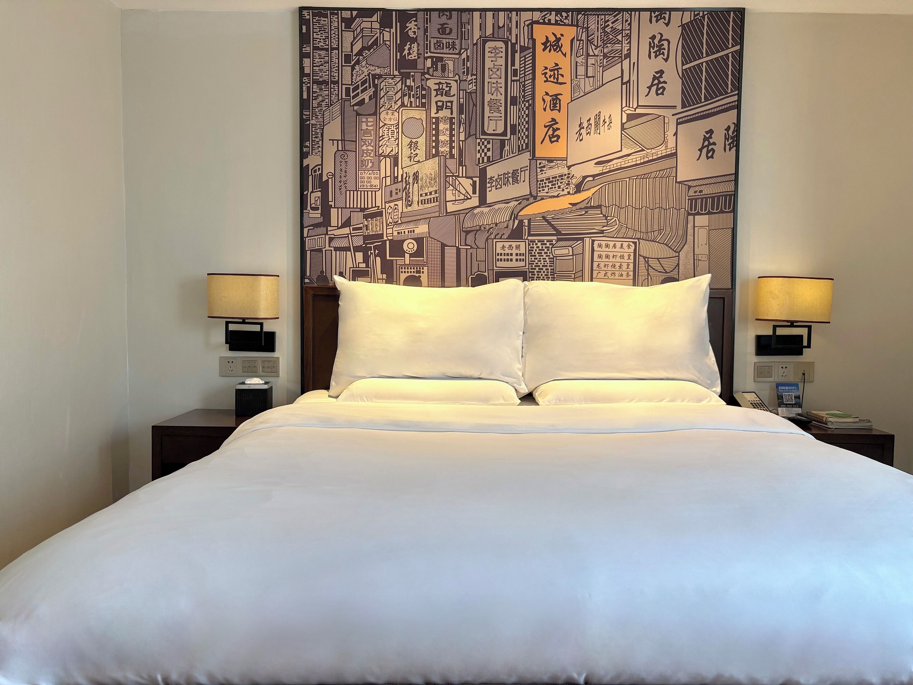 A hotel room with a large bed, two bedside tables with lamps, and a wall tapestry depicting a cityscape.