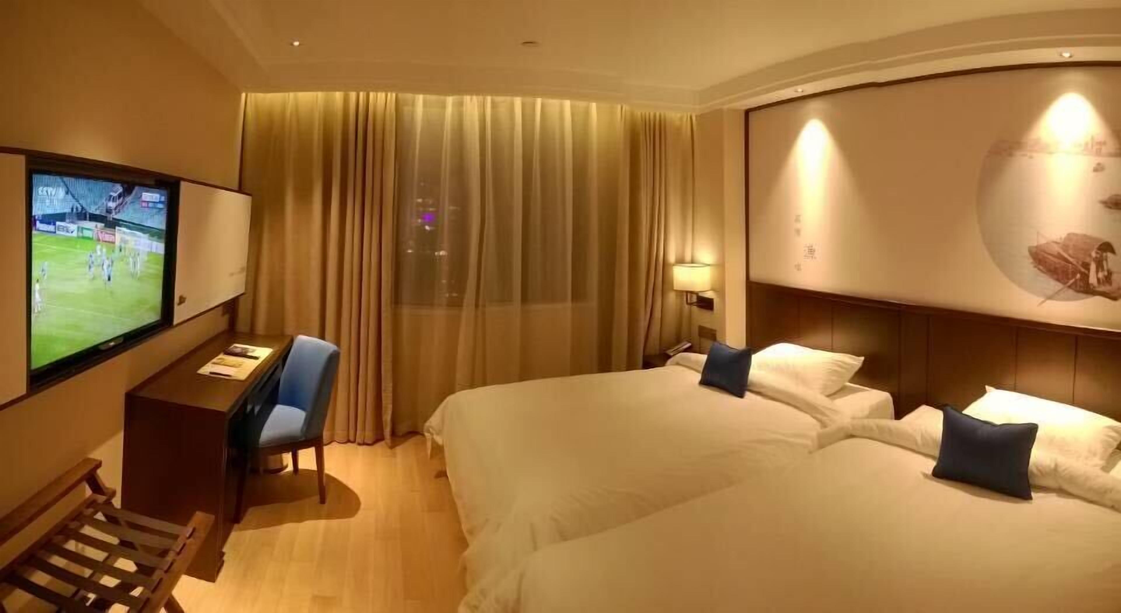 A hotel room with two beds, a desk with a chair, a TV, and a large window with curtains.