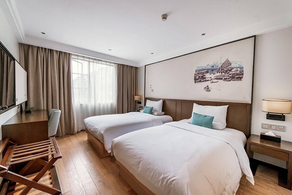 Ein Hotelzimmer mit zwei Betten, einem Schreibtisch, einem Stuhl, einem Fenster mit Vorhängen, einer Wandlampe und einem Gemälde.