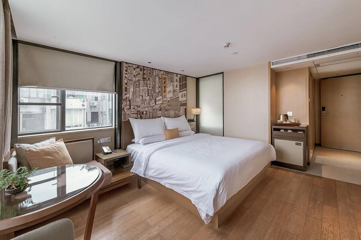 Ein modernes Hotelzimmer mit einem großen Bett, einem Nachttisch mit Lampe, einem kleinen Schreibtisch mit Stuhl und einem Fenster mit Blick auf die Stadt.