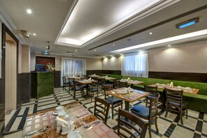 Une salle à manger d’hôtel avec un sol à carreaux, de nombreuses tables et chaises, un canapé vert et un comptoir de réception.