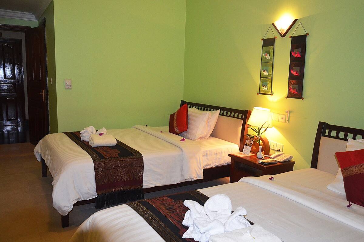 Habitación de hotel con dos camas, una mesita de noche con lámpara, y un estante en la pared con objetos decorativos.