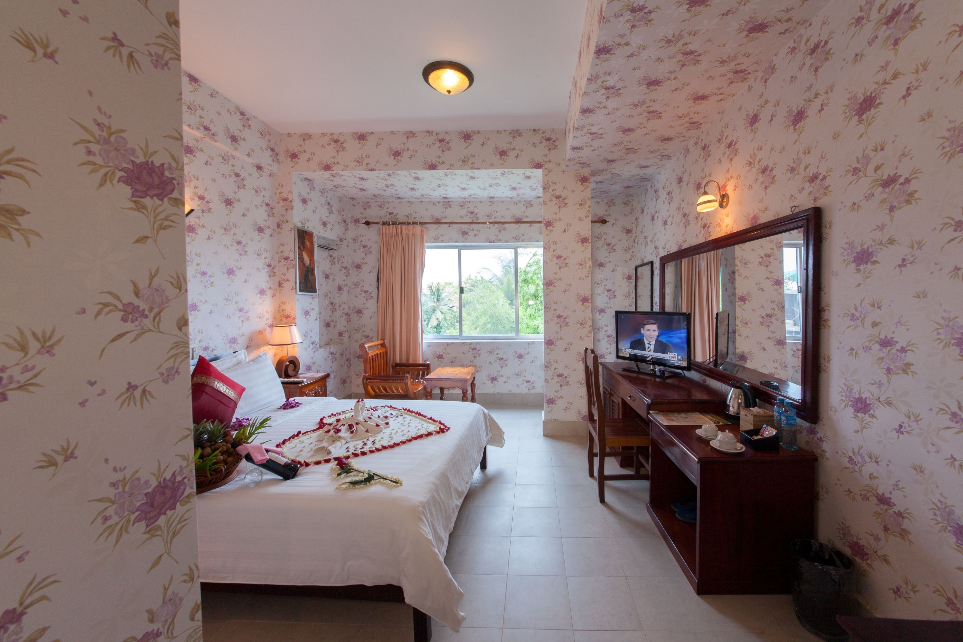 A hotel room with floral wallpaper, a bed with a rose petal arrangement, a desk with a television, and a window with a view of greenery.