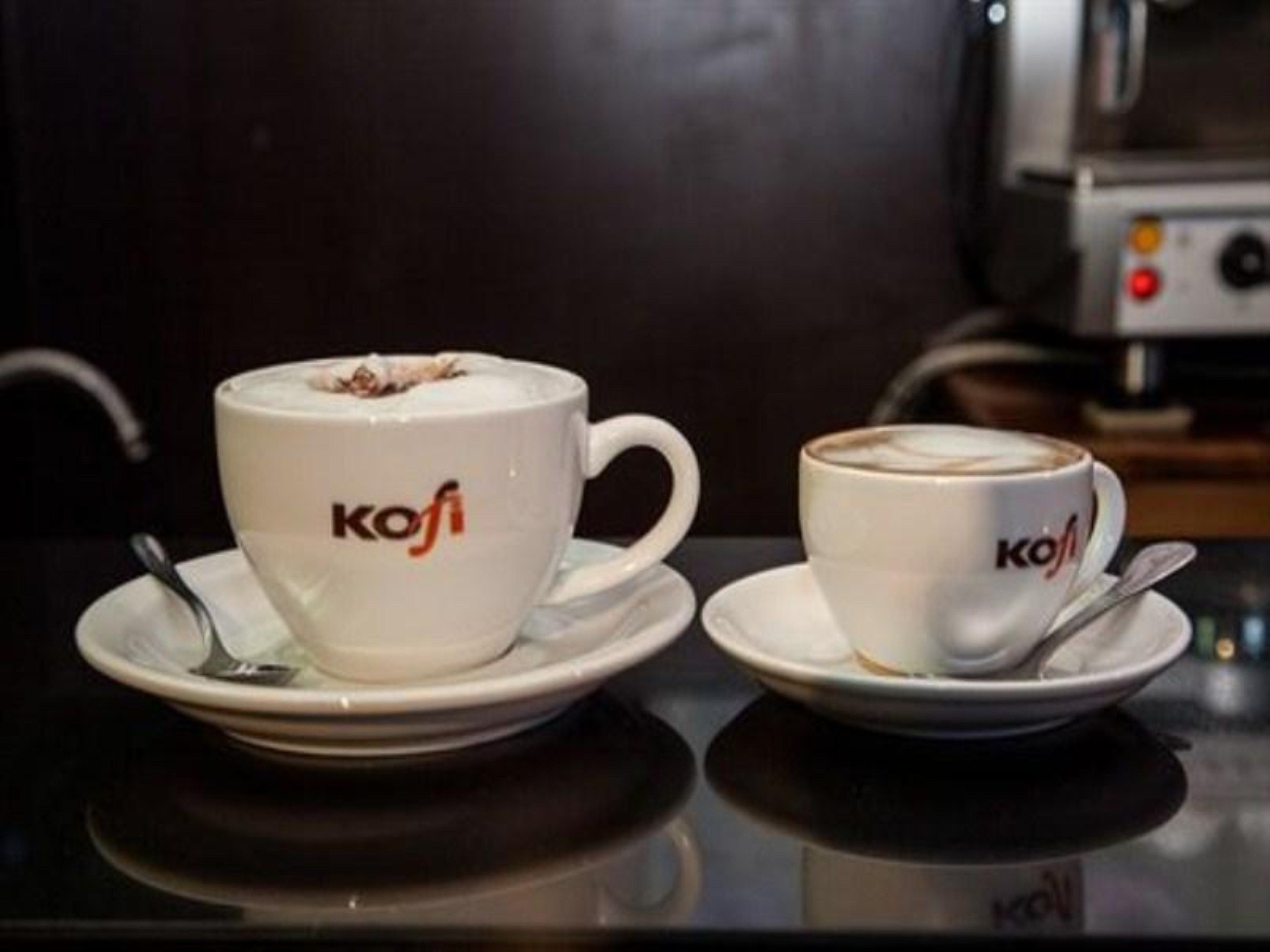 Two white coffee cups with the logo "Kofi" on a reflective surface, accompanied by saucers and spoons.