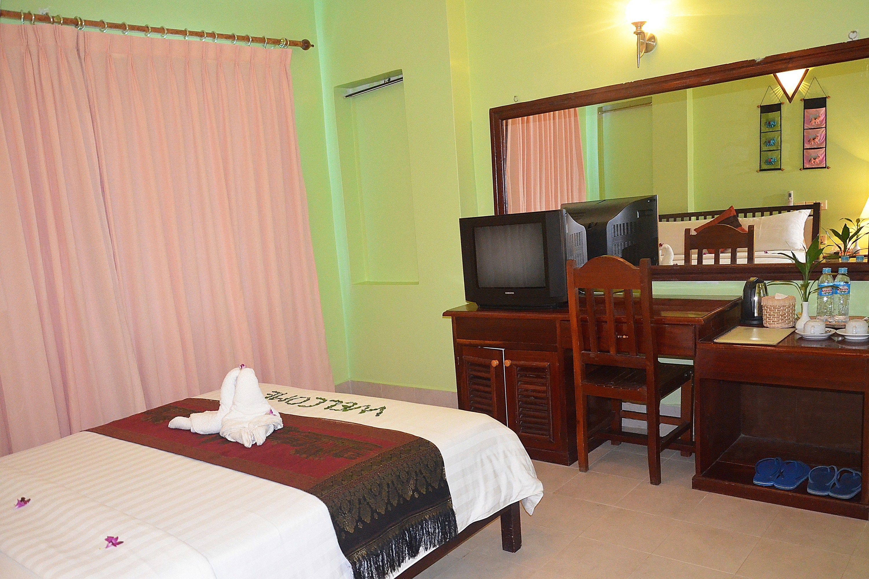 A hotel room with a bed, a desk with a television, a chair, a mirror, and a small table with items on it.