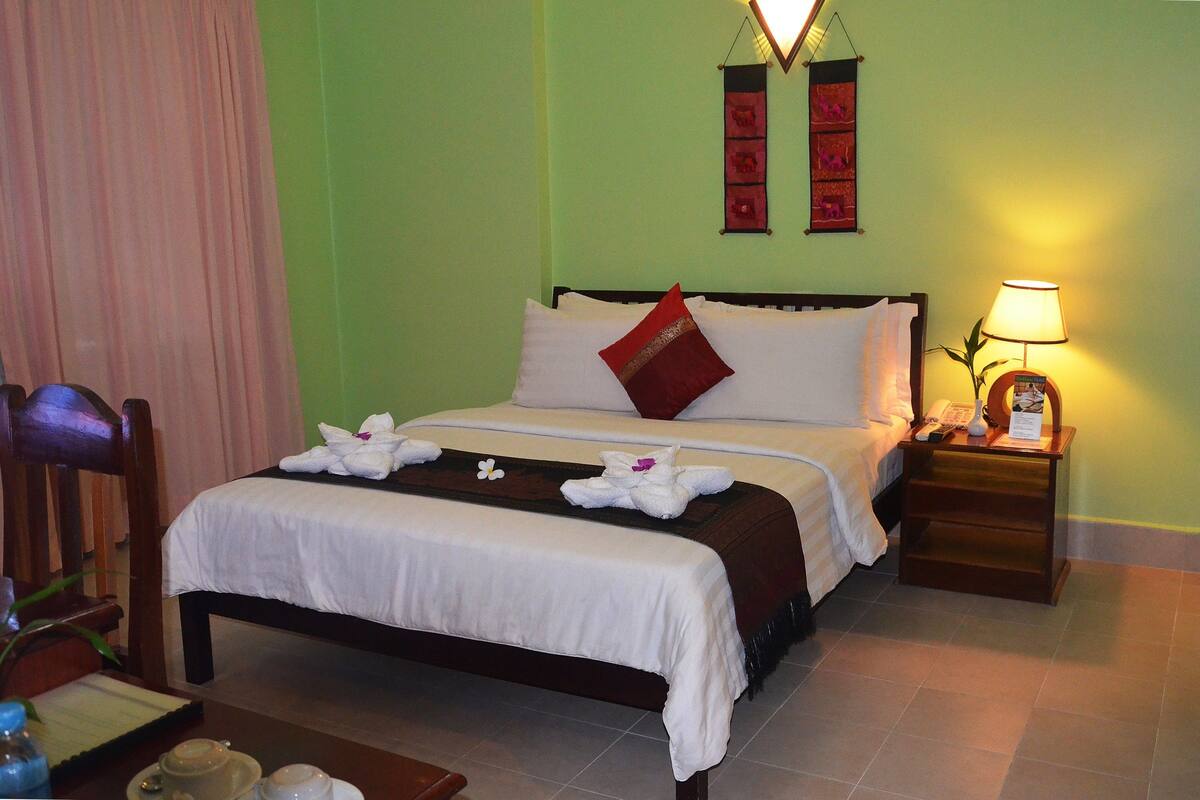 Una cama bien tendida con sábanas blancas y una almohada roja, una mesita de noche con una lámpara, y una silla.
