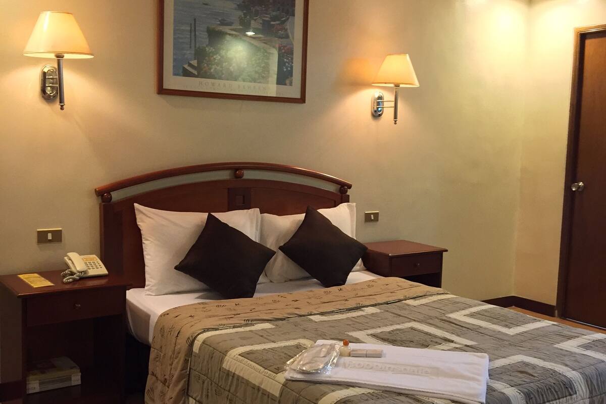 A hotel room with a bed, two bedside lamps, a framed picture on the wall, a telephone on the nightstand, and a door.