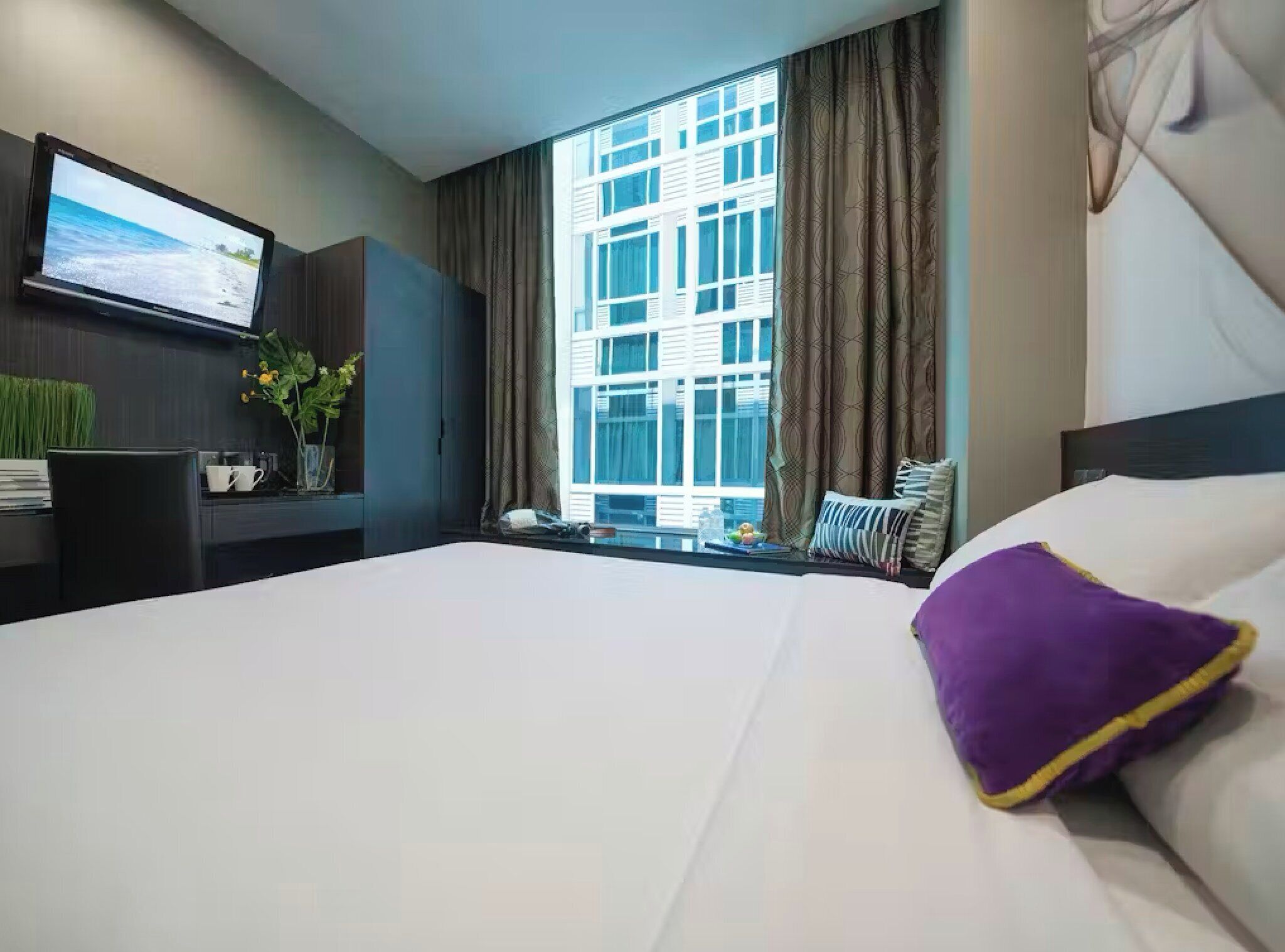 A hotel room with a large bed, a flat-screen TV mounted on the wall, a desk with a chair, and a window with curtains.
