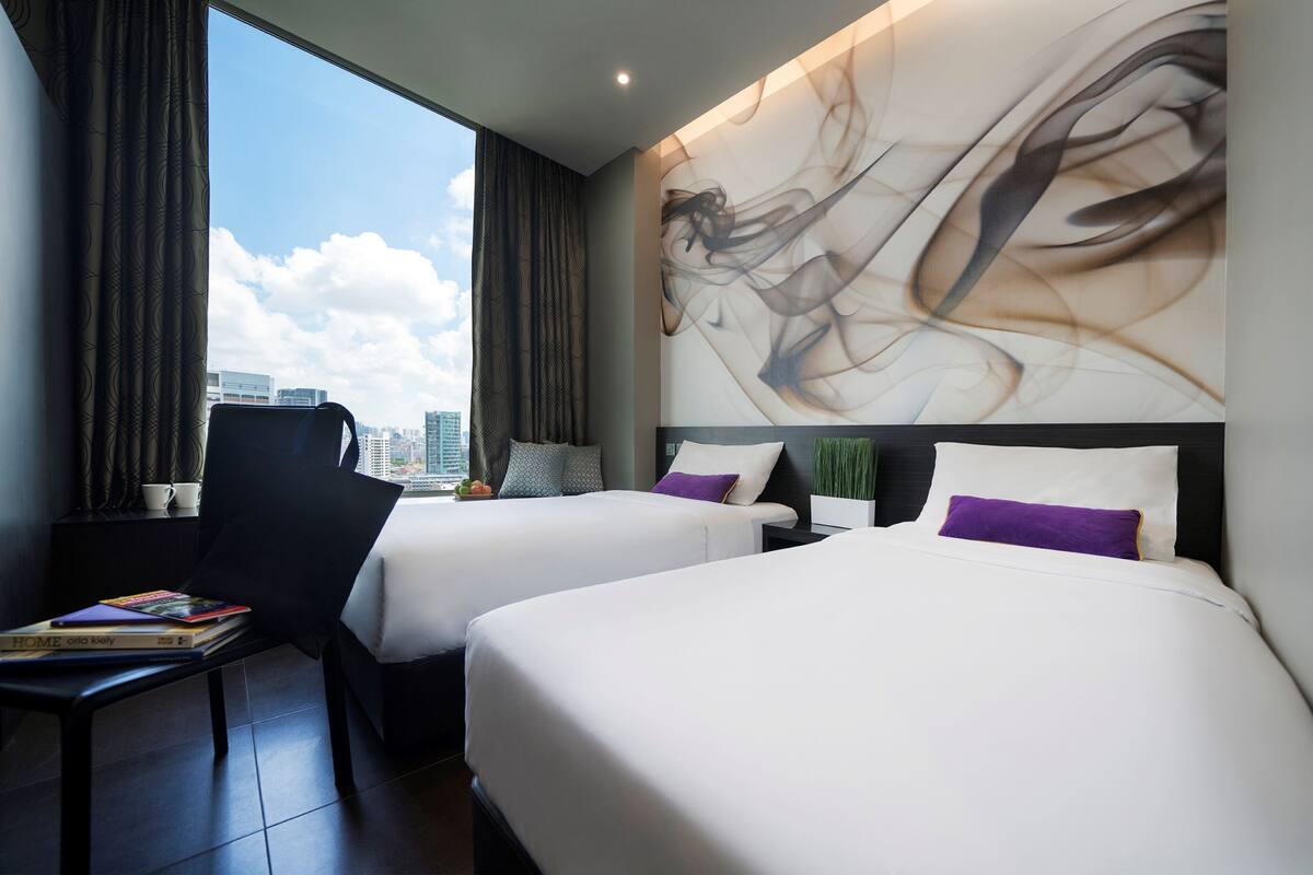 Quarto de hotel com duas camas, uma poltrona, uma mesa e um quadro abstrato grande na parede.