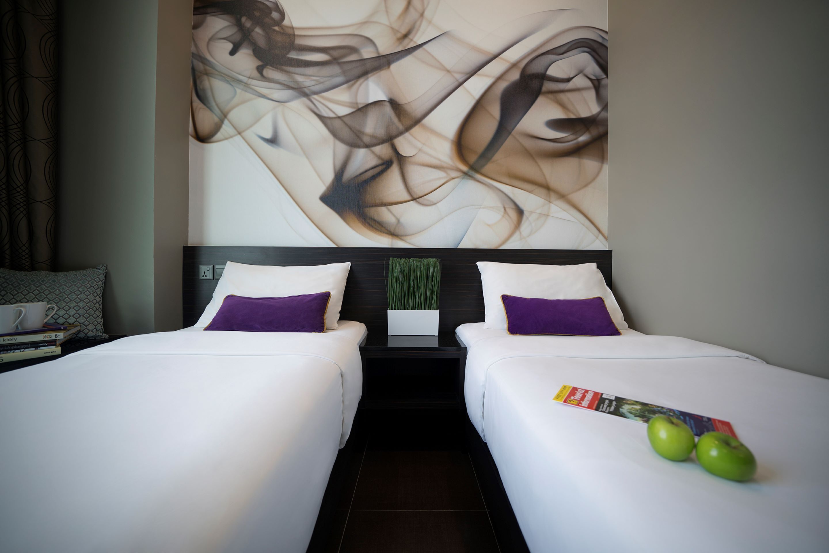 A hotel room with two beds, a large abstract painting on the wall, a small table with a plant, and two apples on the bed.