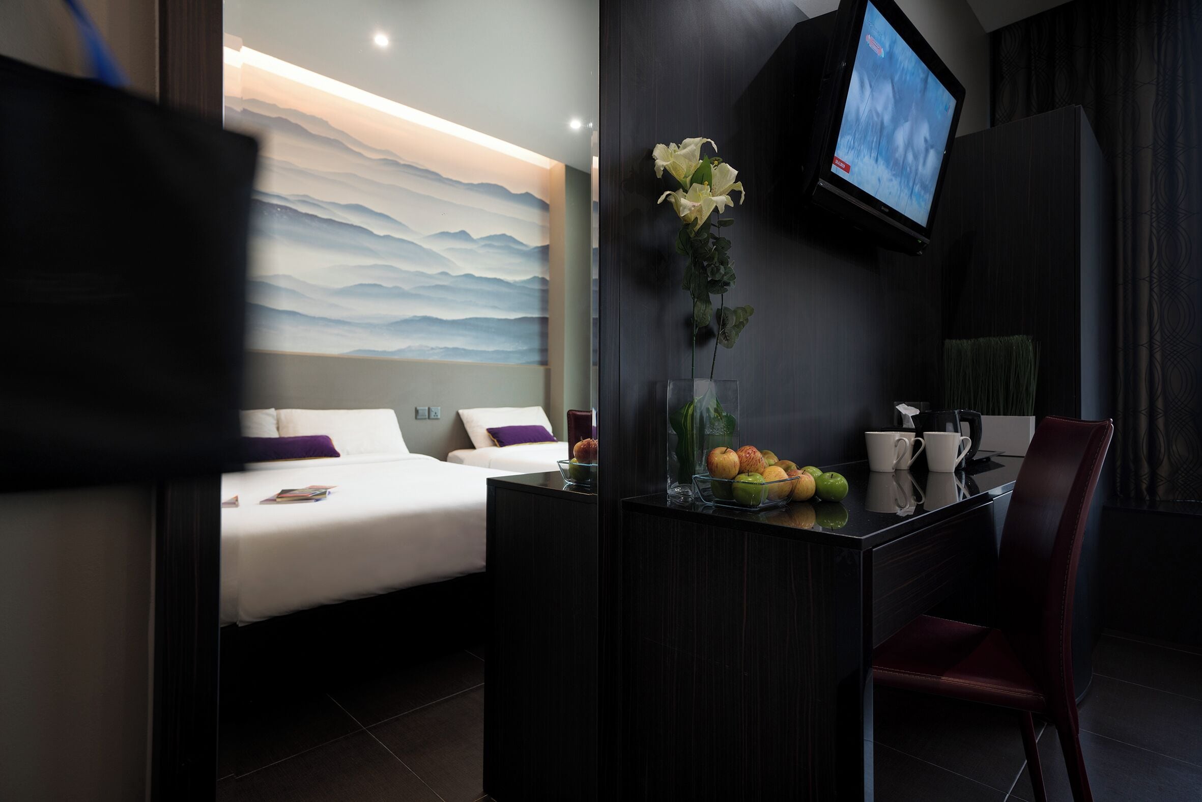 A modern hotel room with a bed, a desk with a vase of flowers, a television, and a view of the mountains.
