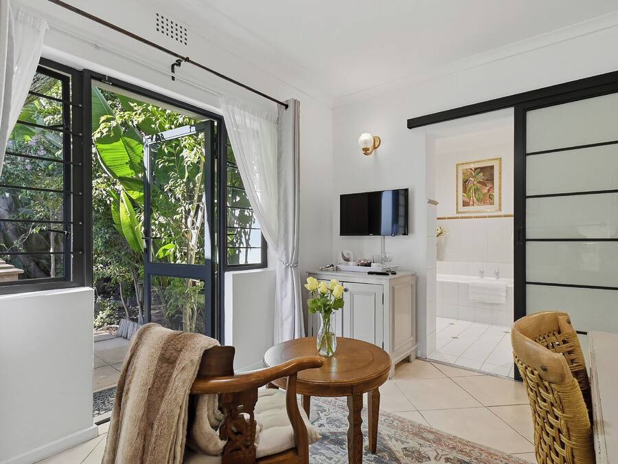 A room with a television, a dining table, chairs, a window with curtains, and a view of greenery outside.