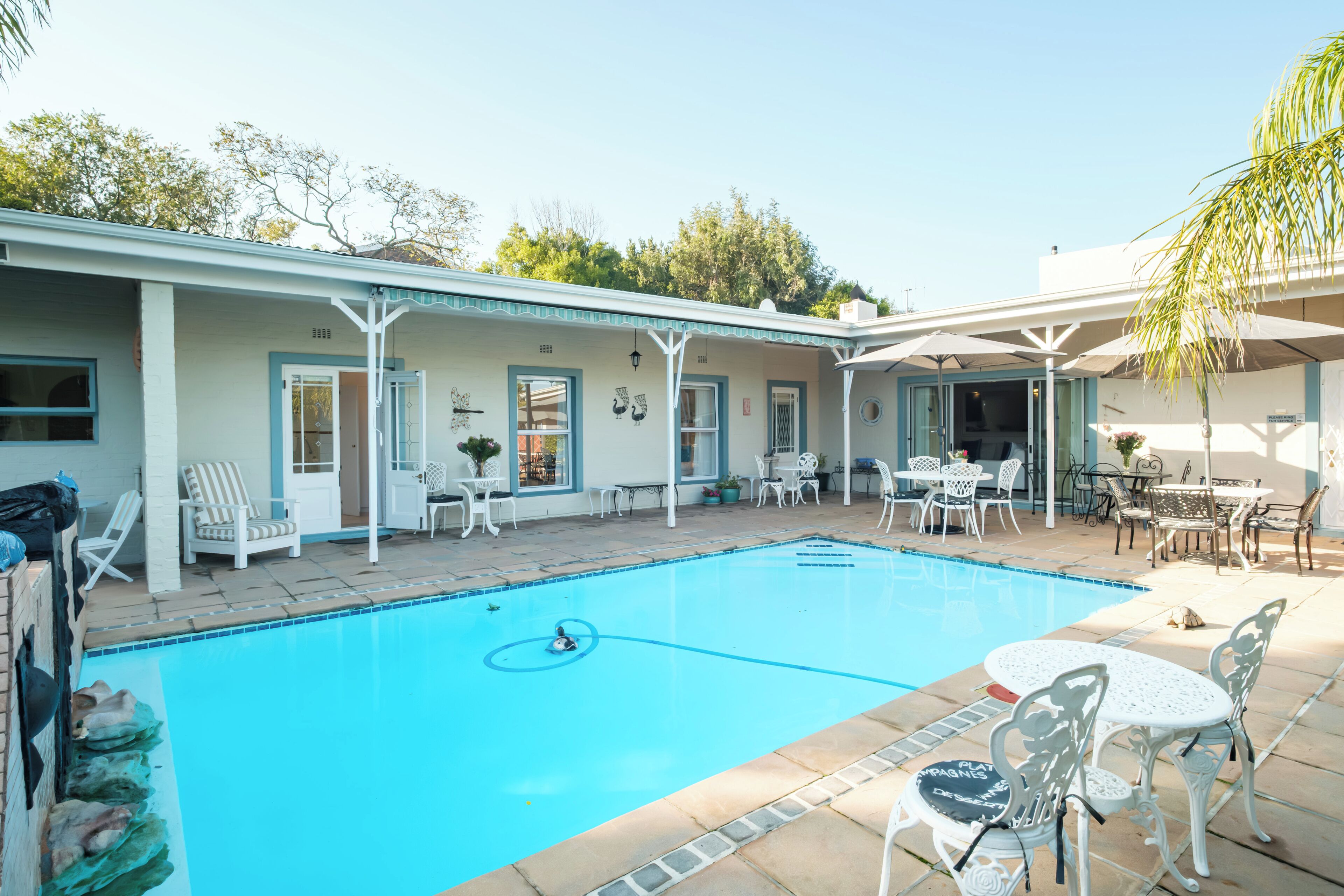 A swimming pool with a sunken center, surrounded by white outdoor furniture, and a covered patio area with tables and chairs.