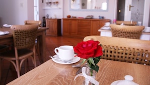 Et spiseområde med fletstole, et træbord, en hvid kop og underkop samt en vase med en rød rose.
