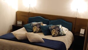 Een net opgemaakt bed met gestreept beddengoed en meerdere kussens, waaronder exemplaren met een tropisch thema. Twee nachtkastjes met lampen staan ernaast, en op één nachtkastje ligt een klein bordje. De kamer heeft een houten vloer en een stoel in de voorgrond.