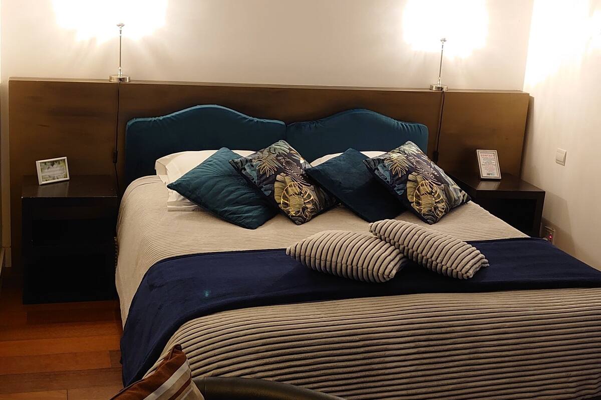 Een net opgemaakt bed met een gestreept dekbed, twee nachtkastjes met lampen en een enkele stoel.
