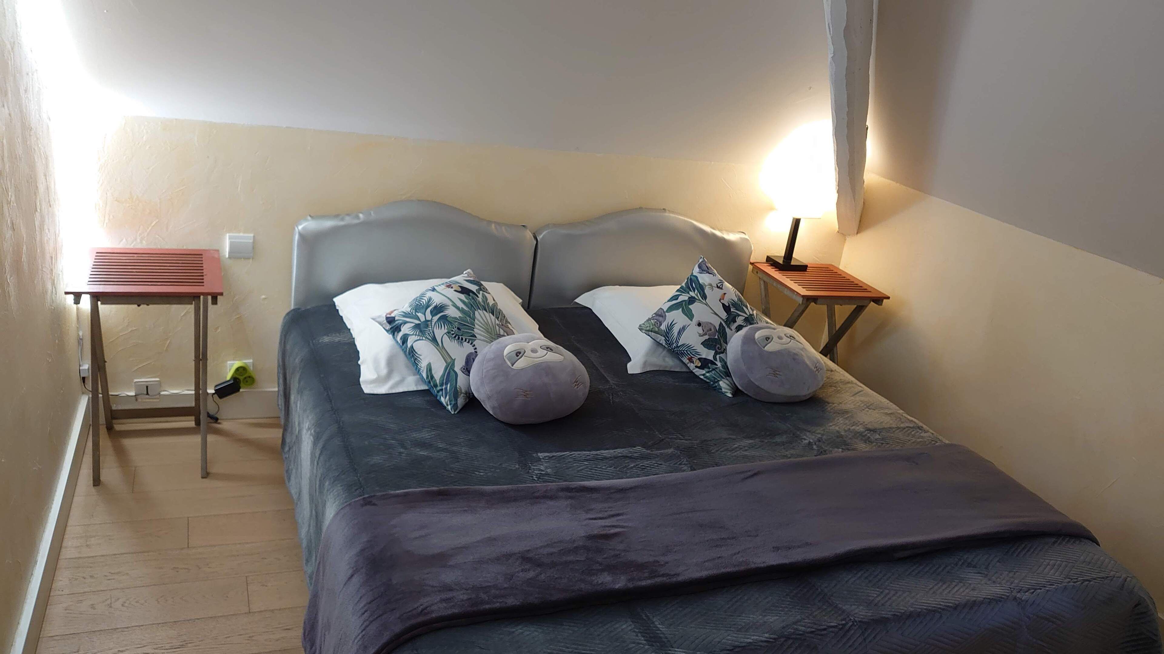 A neatly made bed with a dark blanket, two pillows with a tropical print, and a bedside table with a lamp.