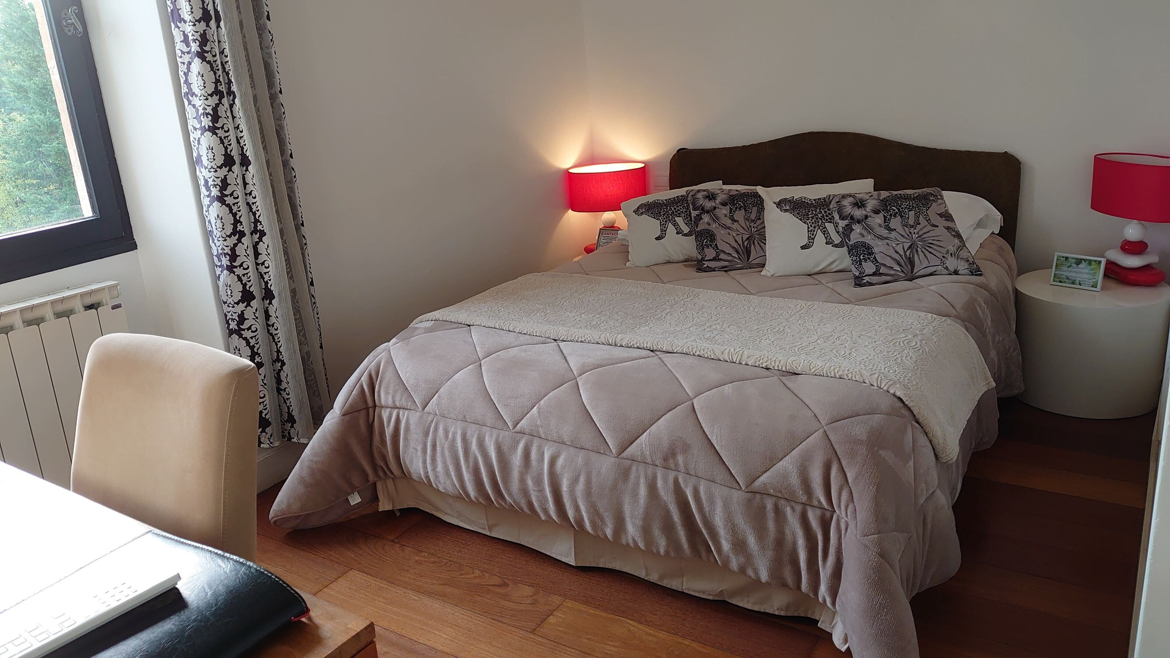 En pænt redt seng med et quiltet betræk, to puder med leopardprint, en rød lampe på sengebordet og et lille bord med en rød genstand på.