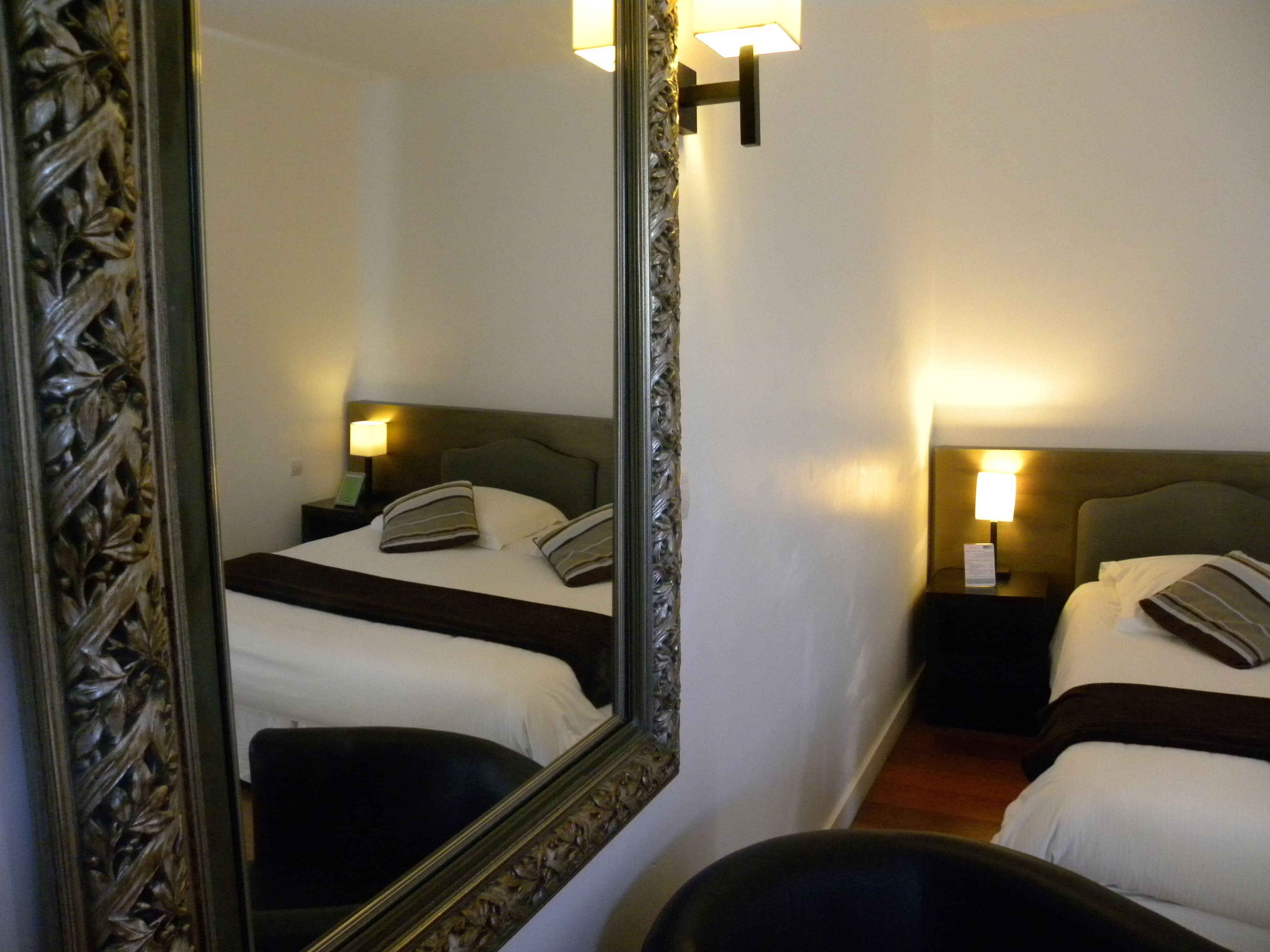 Habitación de hotel con un espejo grande y decorativo, dos camas con sábanas blancas, cabeceros oscuros y lámparas de noche.