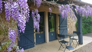 Een gebouw met een stenen gevel, twee donkere houten deuren en een pergola overwoekerd met lila druivenbloemen. Buiten staan twee zwarte stoelen en een kleine ronde tafel.