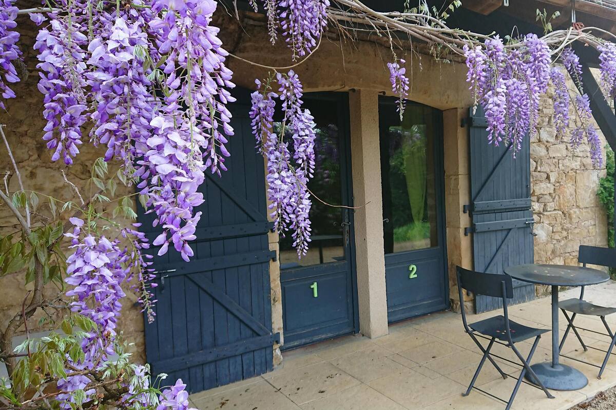 Een gebouw met een stenen gevel, twee donkere houten deuren en een pergola overwoekerd met lila druivenbloemen. Buiten staan twee zwarte stoelen en een kleine ronde tafel.