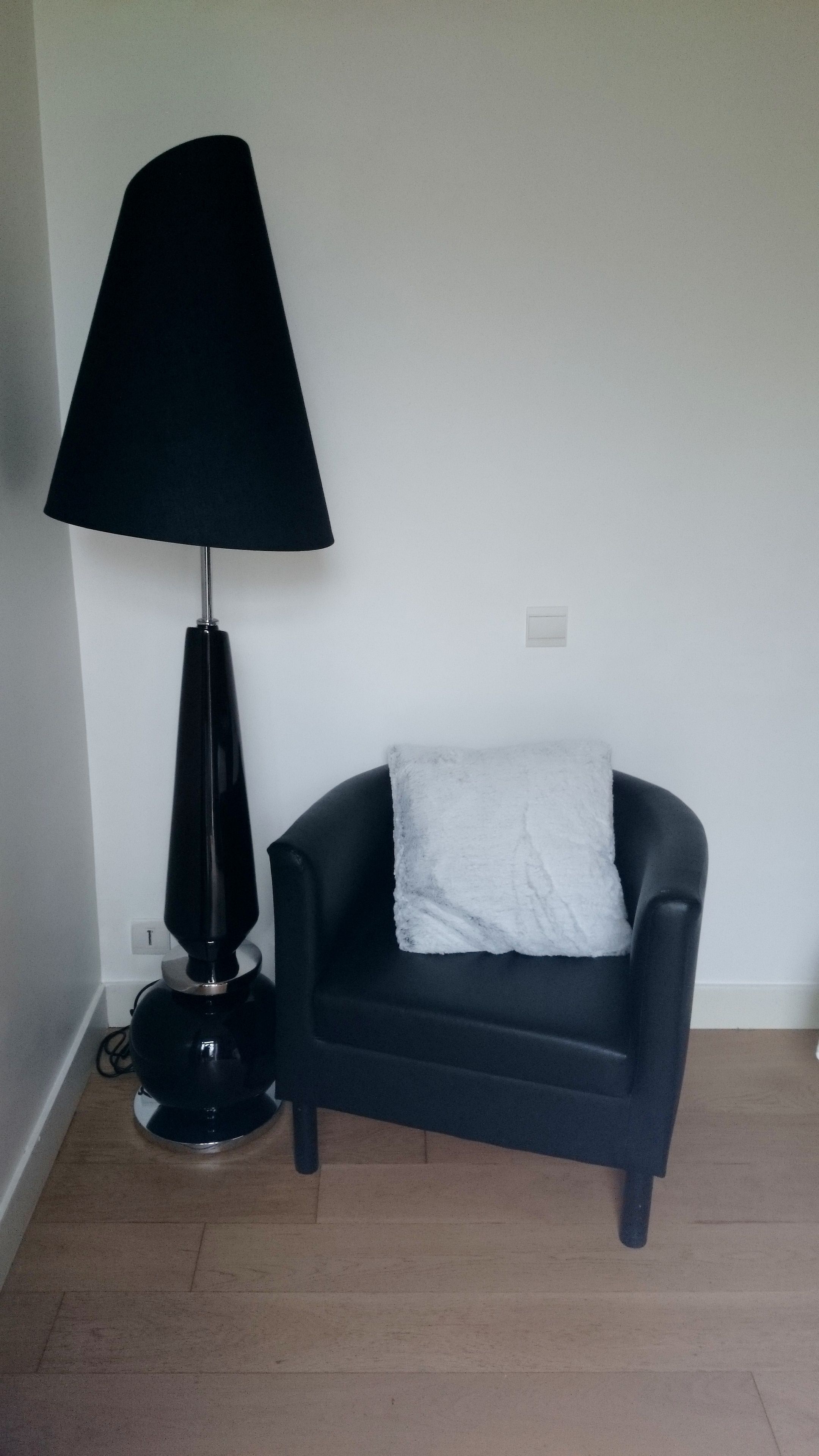 Une pièce d’angle avec un lampadaire noir, un fauteuil sombre agrémenté d’un coussin blanc et un interrupteur sur le mur.