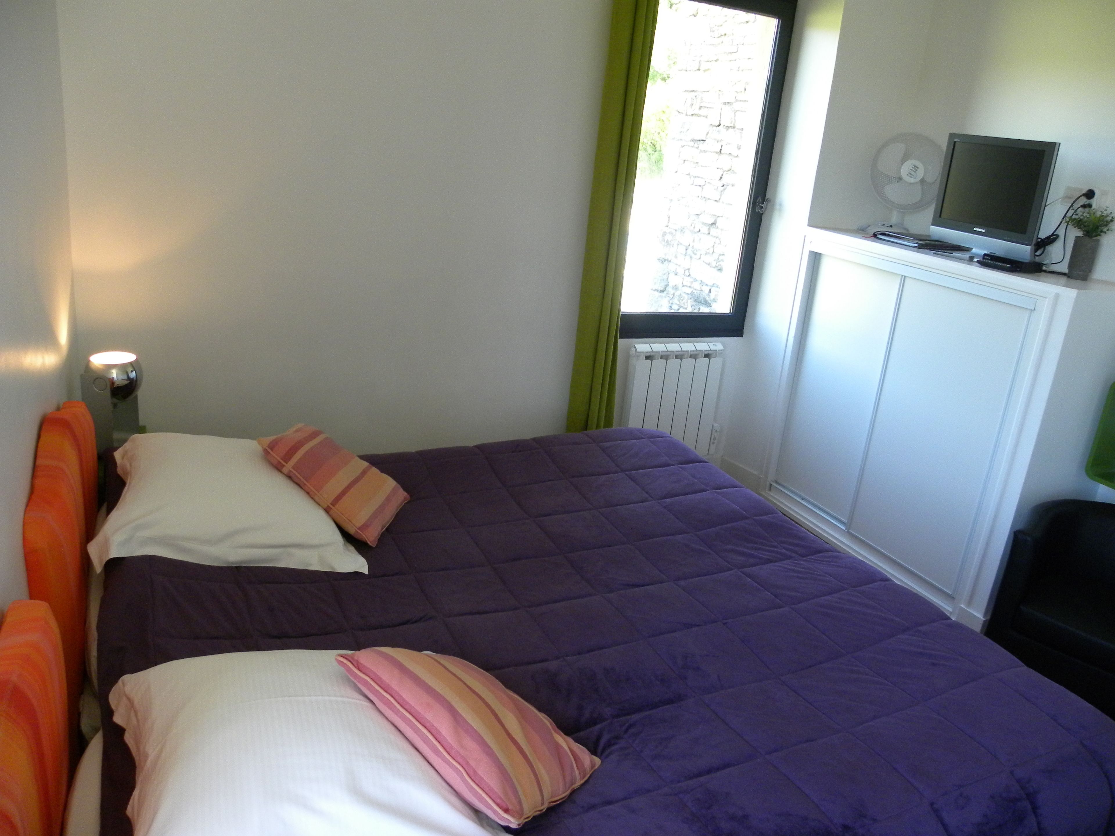 A bedroom with a bed, pillows, a nightstand, a lamp, a radiator, a window with curtains, and a television on a stand.