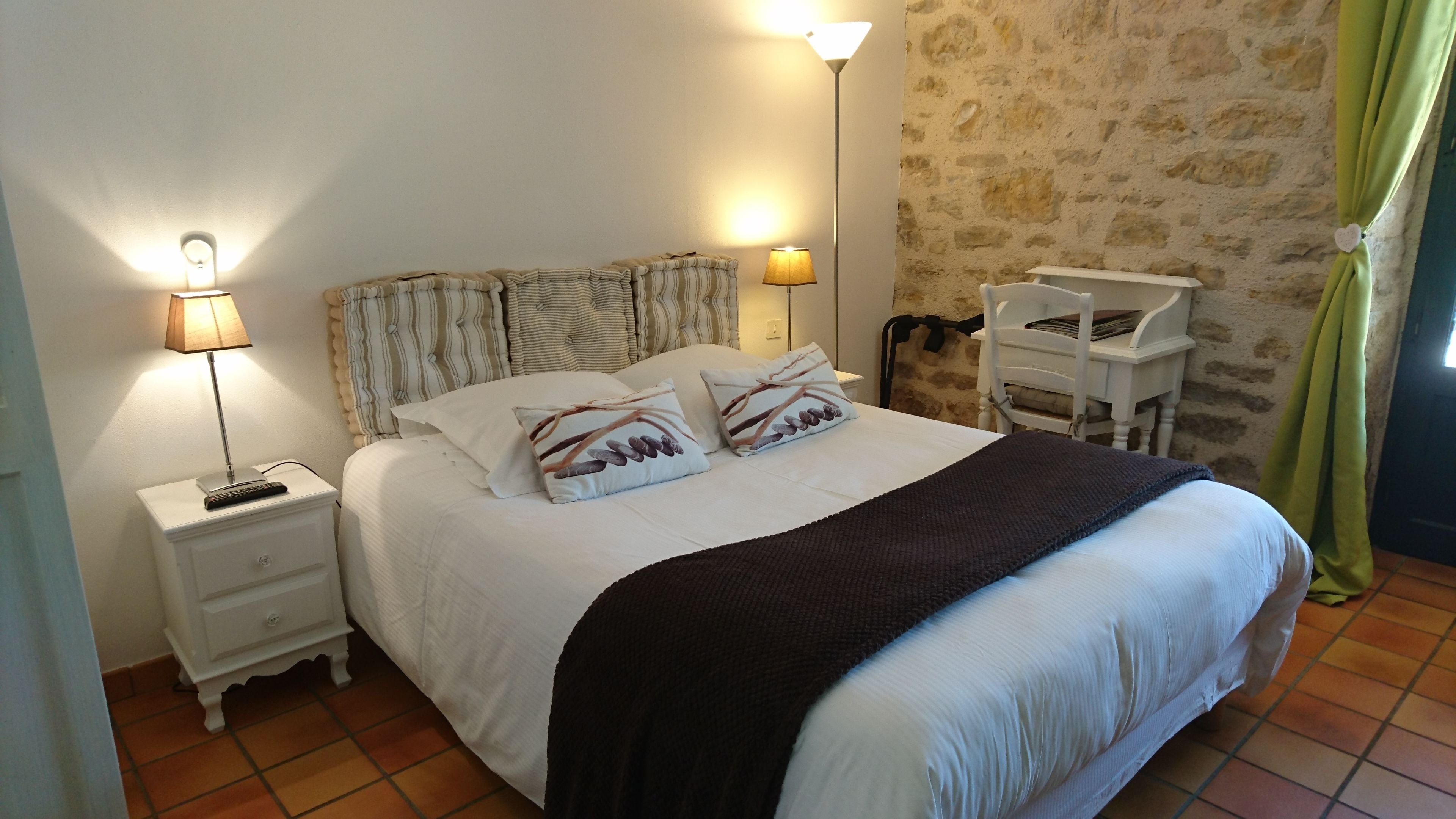 A neatly made bed with striped headboard pillows, a bedside table with a lamp, and a chair against a stone wall.