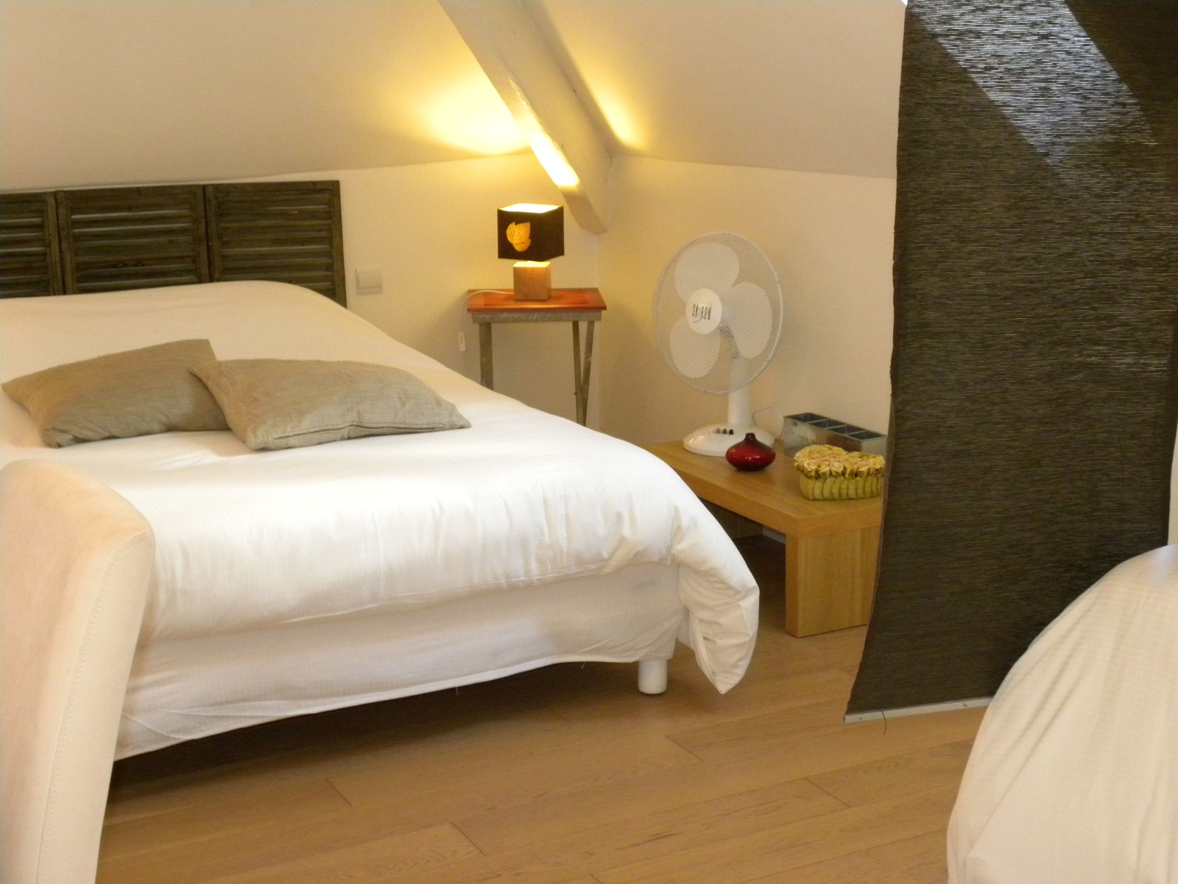 Een net opgemaakt bed met kussens, een nachtkastje met een ventilator en een klein decoratief object.