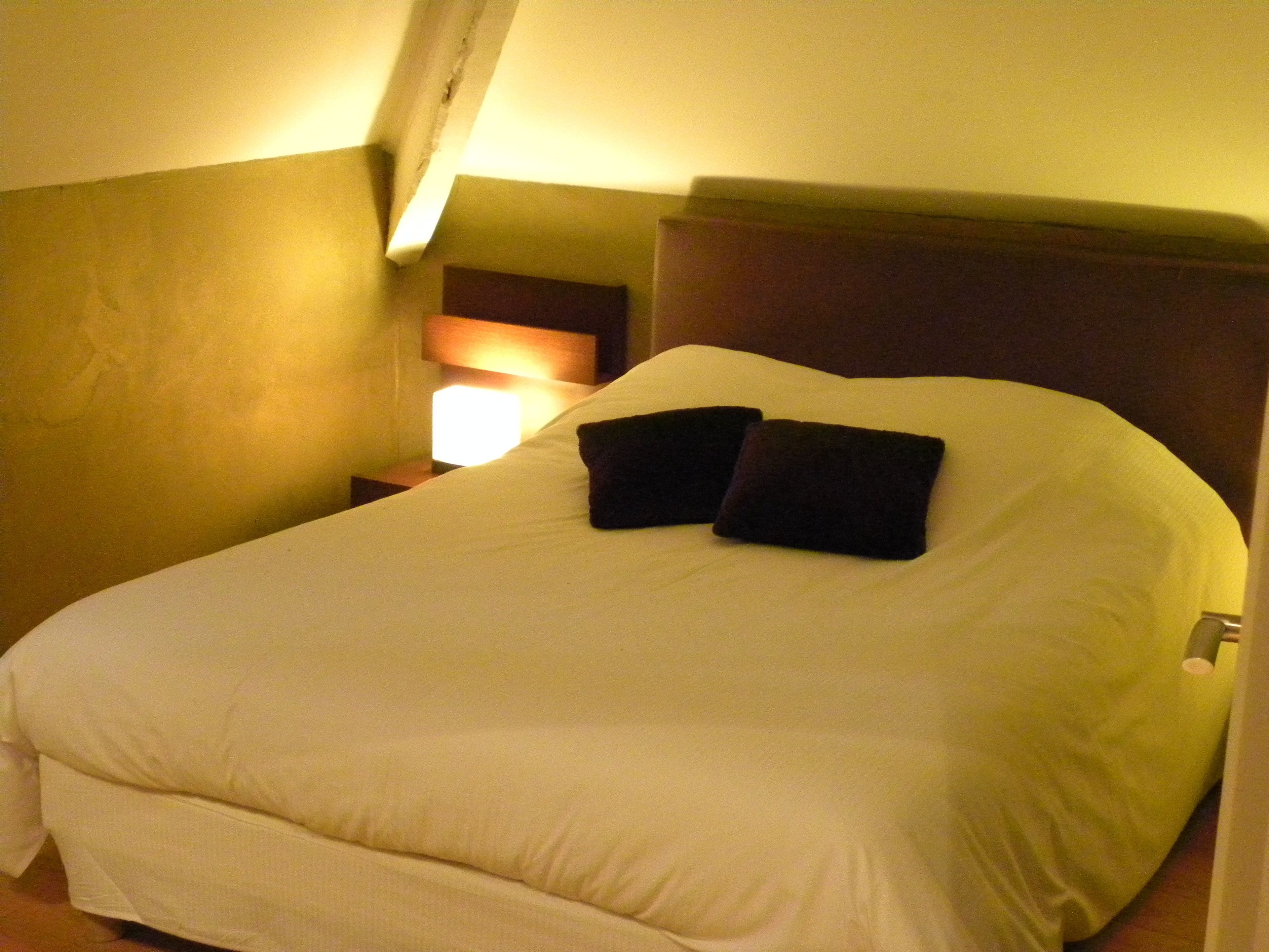 A neatly made bed with white linens and two dark pillows, a bedside lamp, and a wooden headboard in a hotel room.
