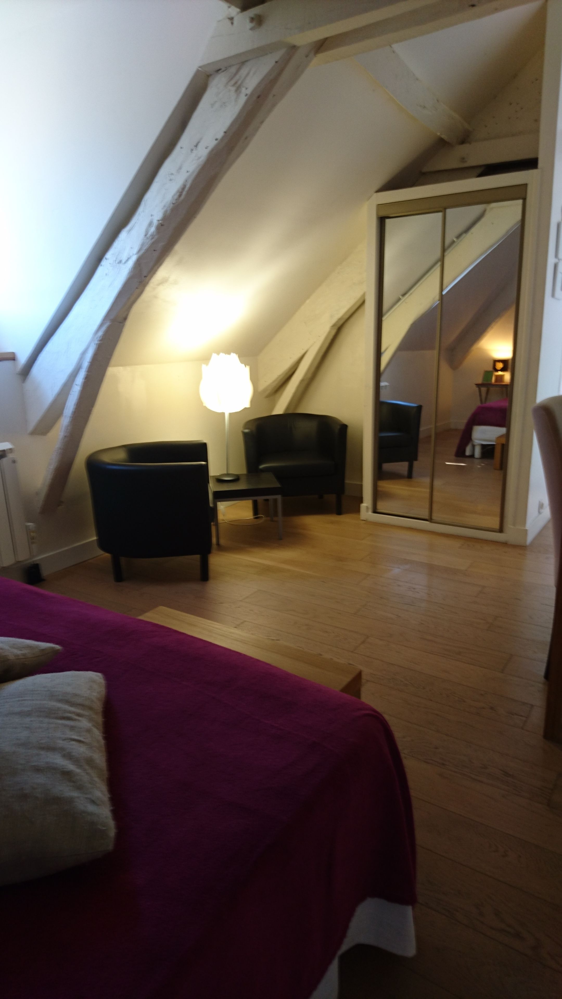 Una habitación con techo inclinado, una cama con colcha roja, una mesita con una silla y una lámpara.