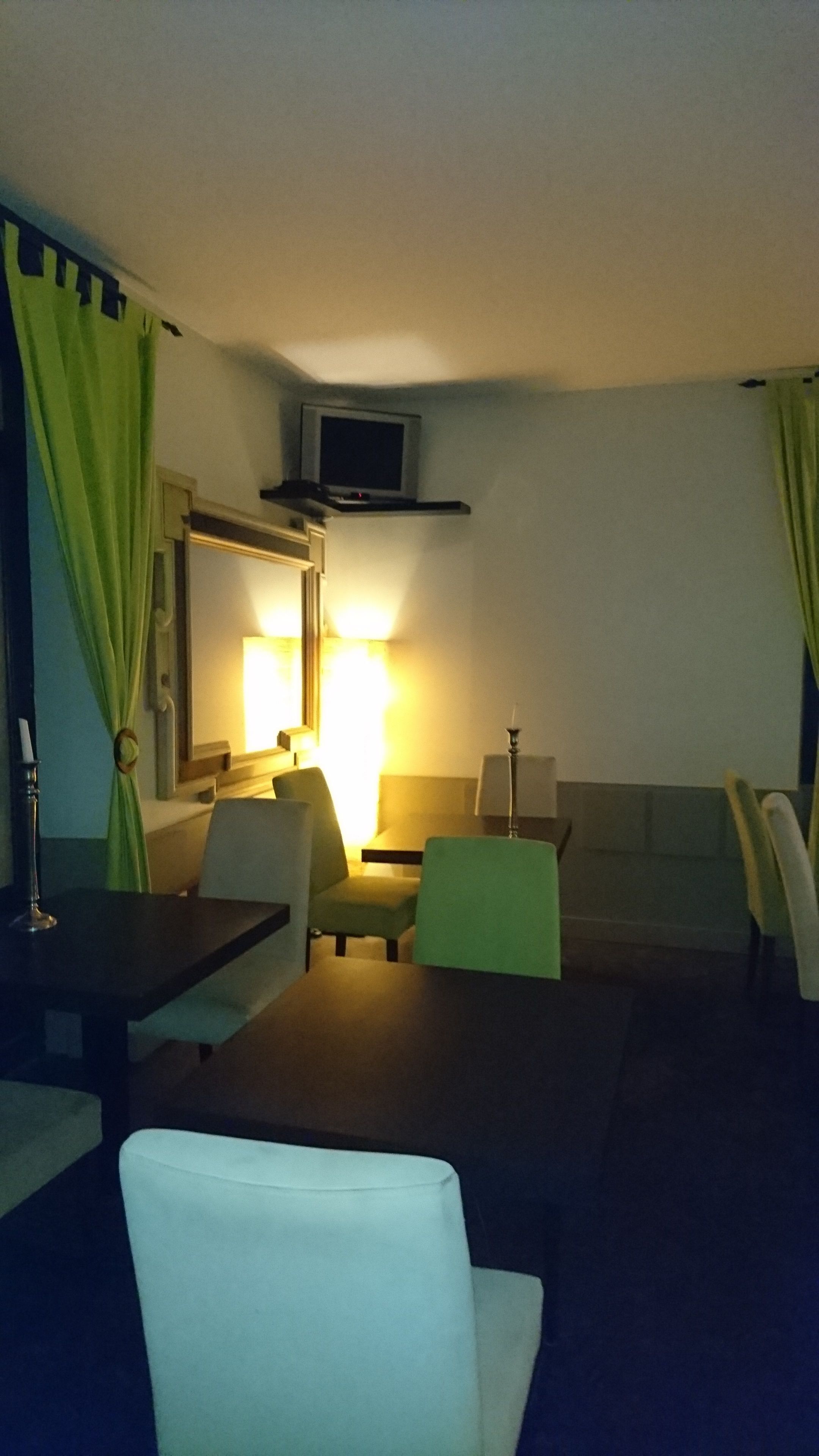 A small, dimly lit room with a single bed, a bedside table, a chair, and a television mounted on the wall.