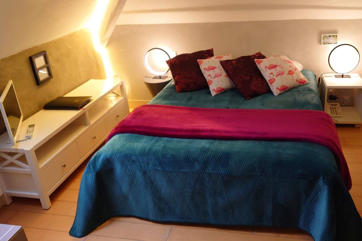 Een slaapkamer met een bed, bedlampen, een bureau met een laptop en een klein plankje.
