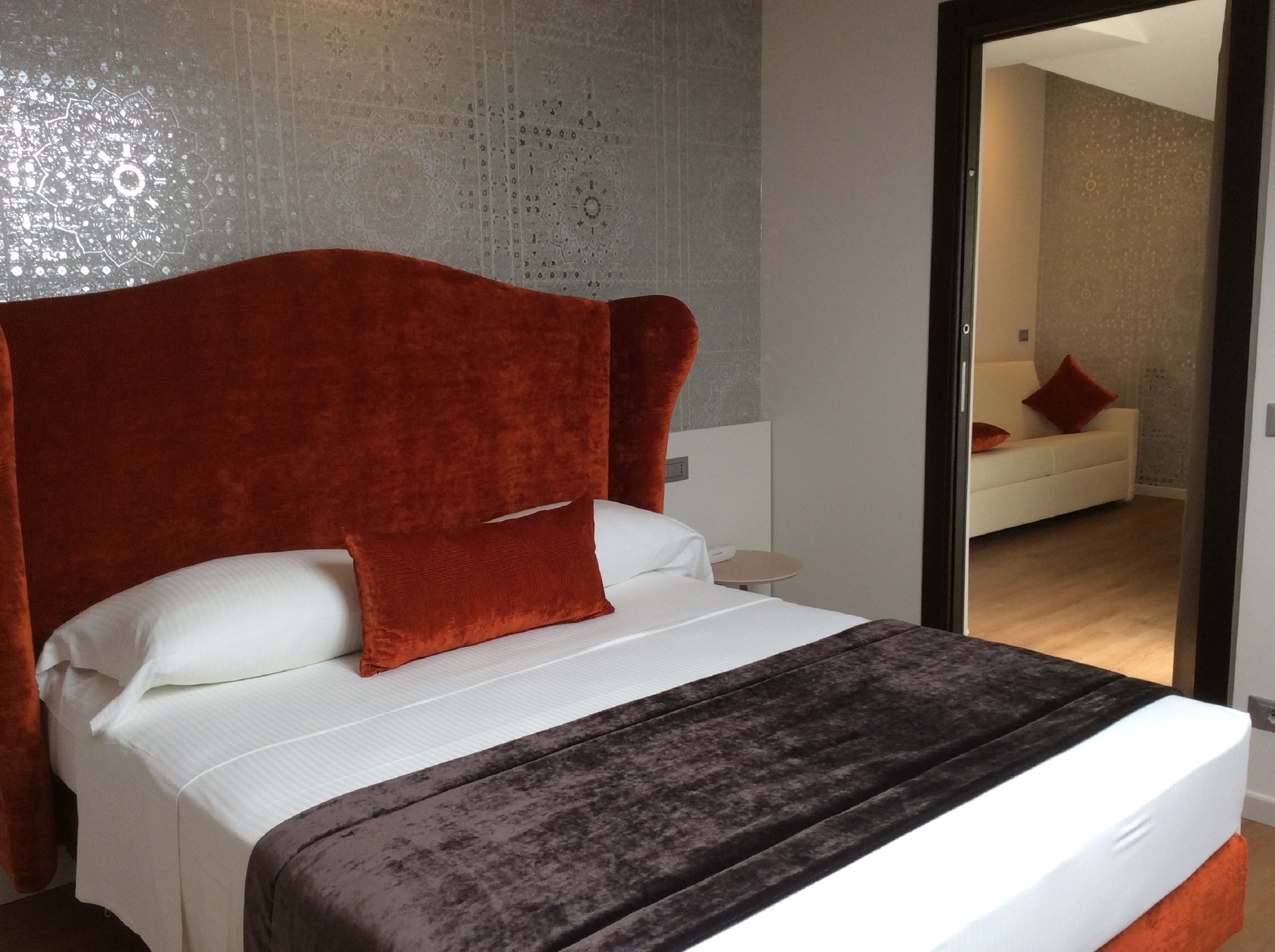 Una moderna camera d'albergo con un letto grande, una testiera rossa e uno specchio che riflette una zona giorno separata con un divano e un cuscino rosso.
