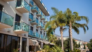 A modern hotel with multiple balconies, large windows, and a view of palm trees and a mountain in the background. - Hotel Ariston (Varazze)