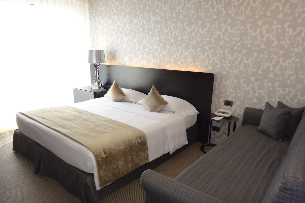 Ein Hotelzimmer mit einem großen Bett, einem Nachttisch mit Lampe, einem kleinen Tisch mit Telefon und einem Sofa.