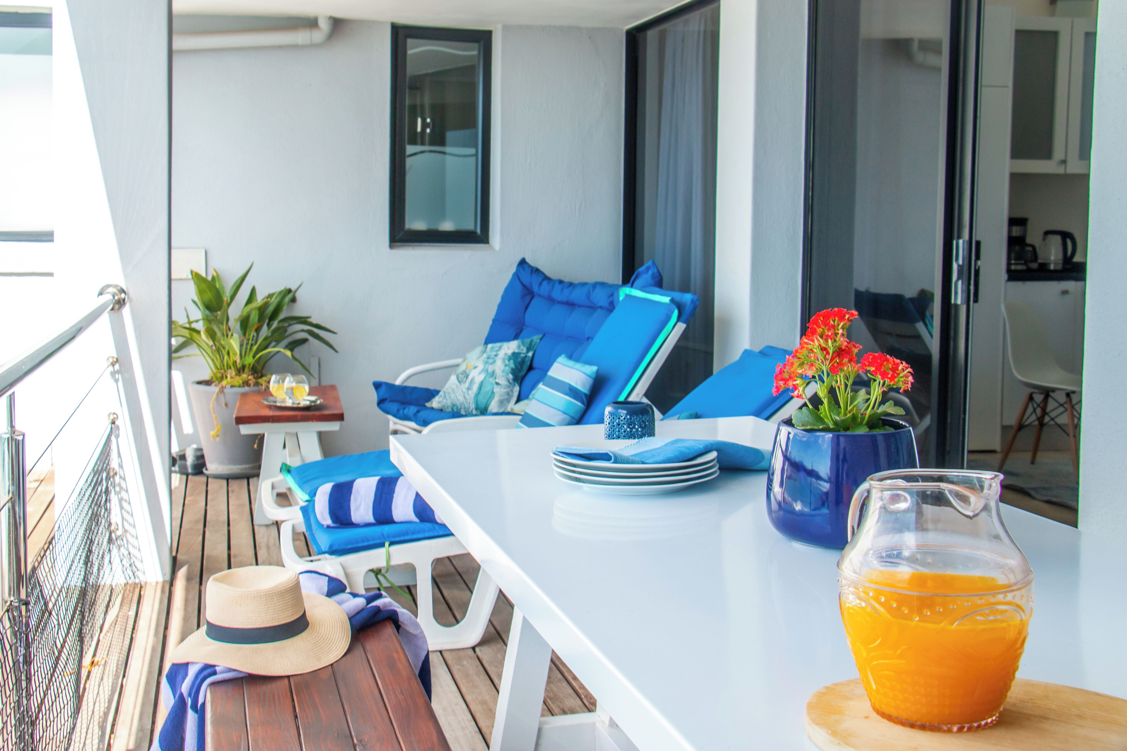 A balcony with a white table, blue cushions, a potted plant, and a glass pitcher of orange juice.