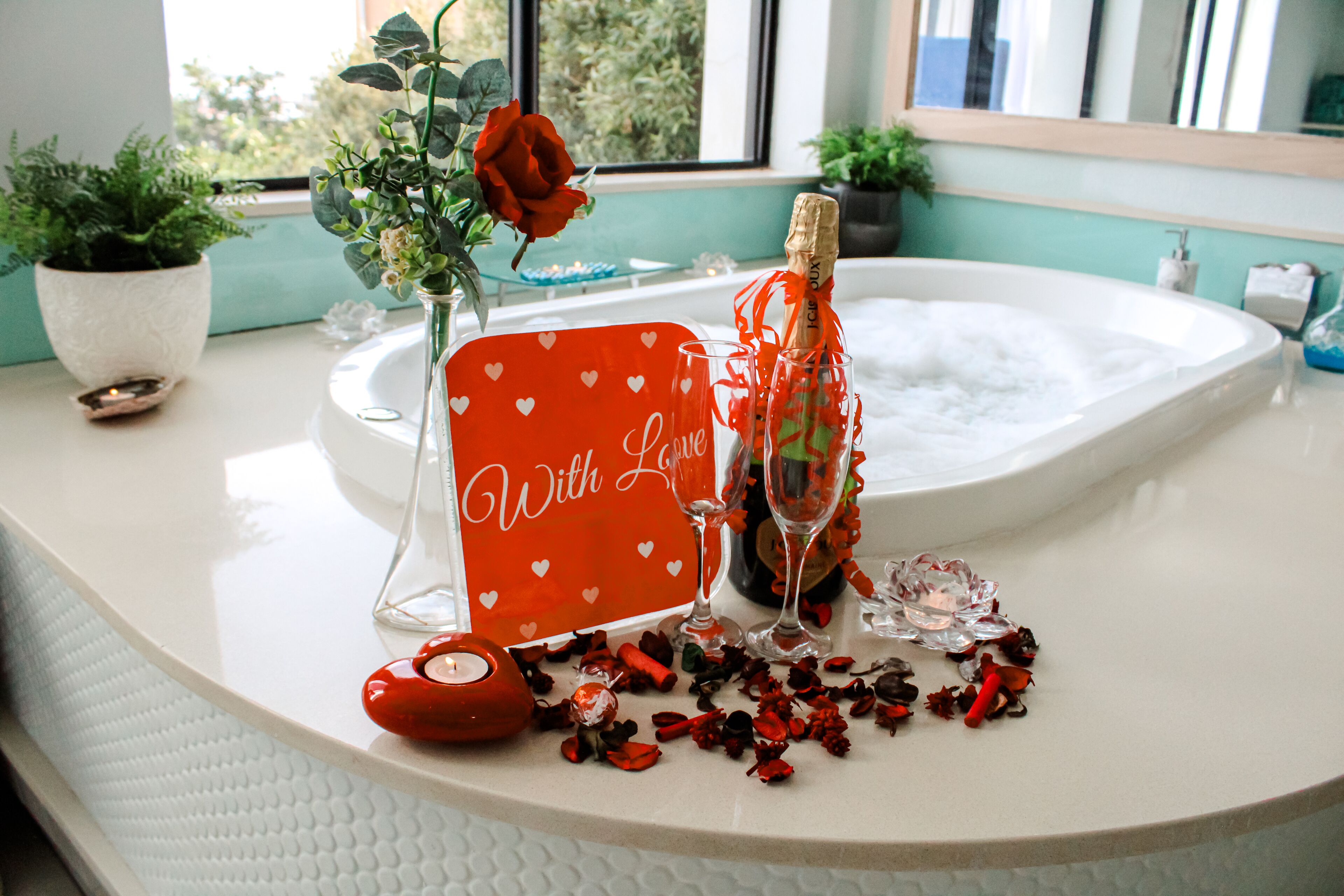Un meuble-lavabo de salle de bain avec un plan de toilette blanc, un panneau rouge orné de cœurs blancs, un vase contenant une rose rouge, une bouteille de champagne et des pétales de rose éparpillés.